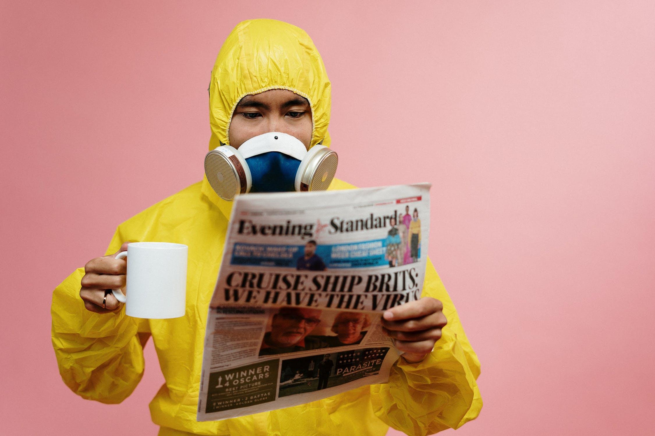 A man with a respirator, reading newspaper and drinking a cup of coffee